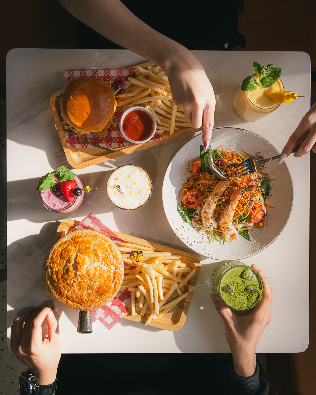 Overhead view of a brunch spread at Odette House Cafe in Spotswood, featuring a beef burger, a meat pie with fries, and a fresh sausage salad being shared by friends.